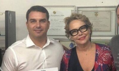 Flávio Bolsonaro e Rogéria Bolsonaro sorriem para foto em ambiente interno, com quadros ao fundo