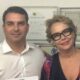 Flávio Bolsonaro e Rogéria Bolsonaro sorriem para foto em ambiente interno, com quadros ao fundo