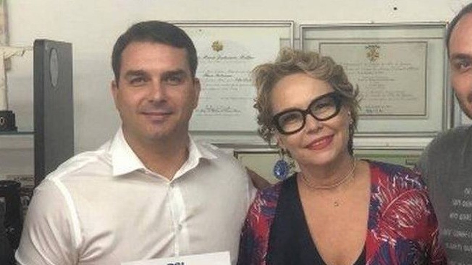 Flávio Bolsonaro e Rogéria Bolsonaro sorriem para foto em ambiente interno, com quadros ao fundo