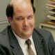 Homem careca, Brian Baumgartner, de terno, sorri maliciosamente em escritório