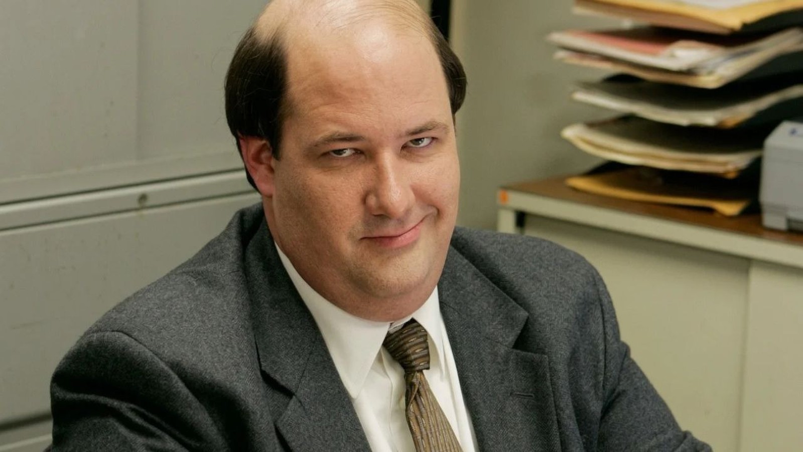 Homem careca, Brian Baumgartner, de terno, sorri maliciosamente em escritório