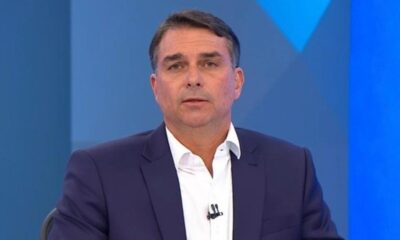 Flávio Bolsonaro em close-up, veste camisa branca e paletó azul escuro. Fundo azul desfocado.