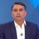 Flávio Bolsonaro em close-up, veste camisa branca e paletó azul escuro. Fundo azul desfocado.