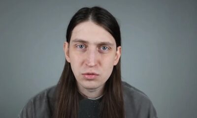 Homem de cabelos longos, olhos claros e expressão neutra olhando para a câmera com microfone