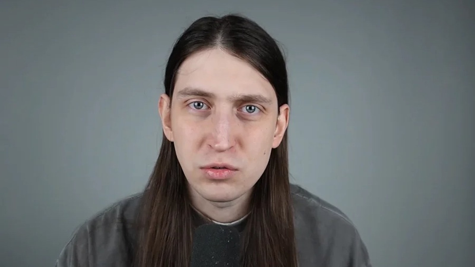Homem de cabelos longos, olhos claros e expressão neutra olhando para a câmera com microfone