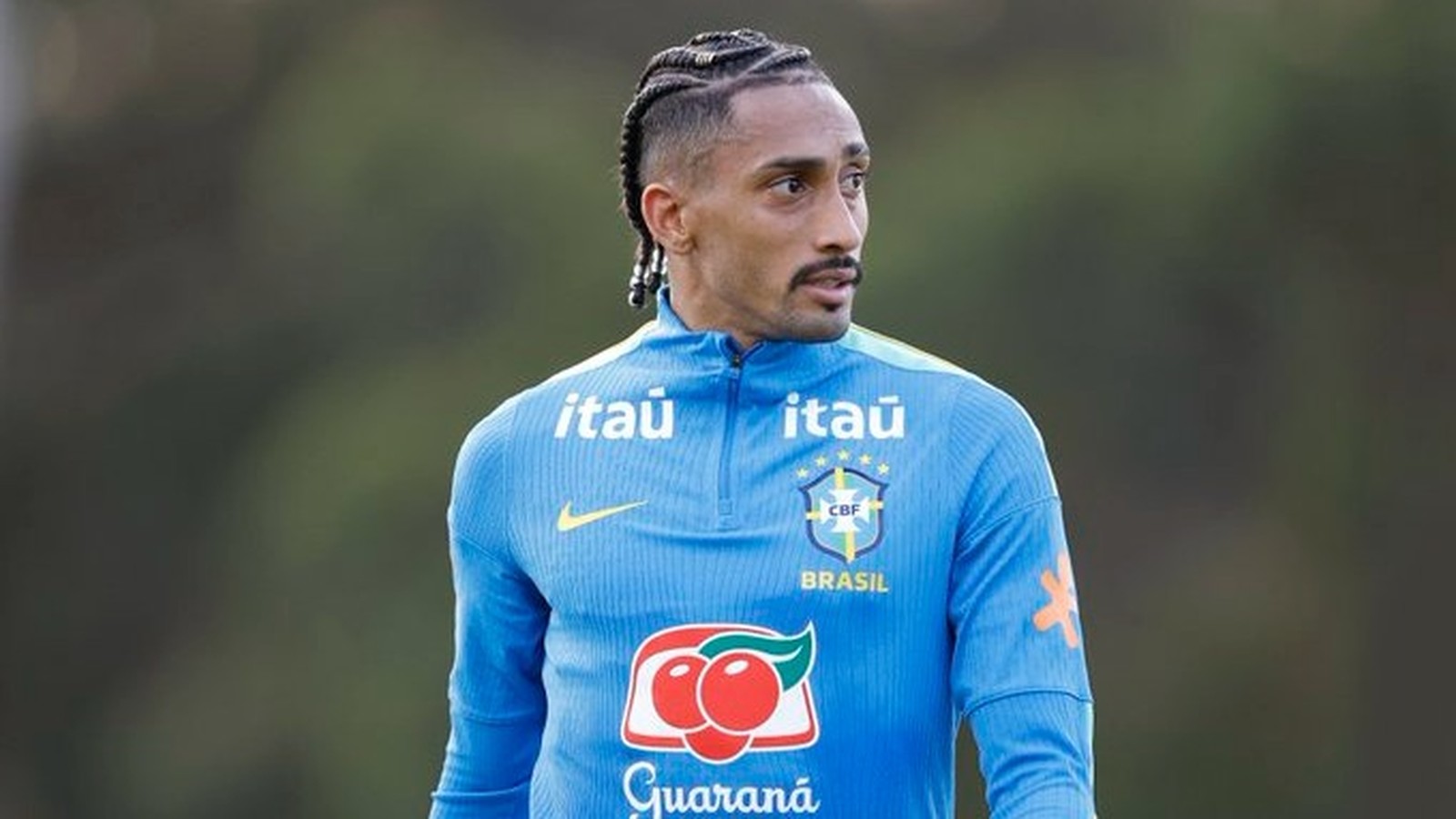 Raphinha, com tranças e bigode, em camisa de treino azul da Seleção Brasileira com logos Itaú e Guaraná