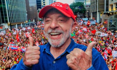 Lula sorrindo, dois polegares para cima, boné vermelho, pin PT, multidão ao fundo