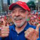 Lula sorrindo, dois polegares para cima, boné vermelho, pin PT, multidão ao fundo