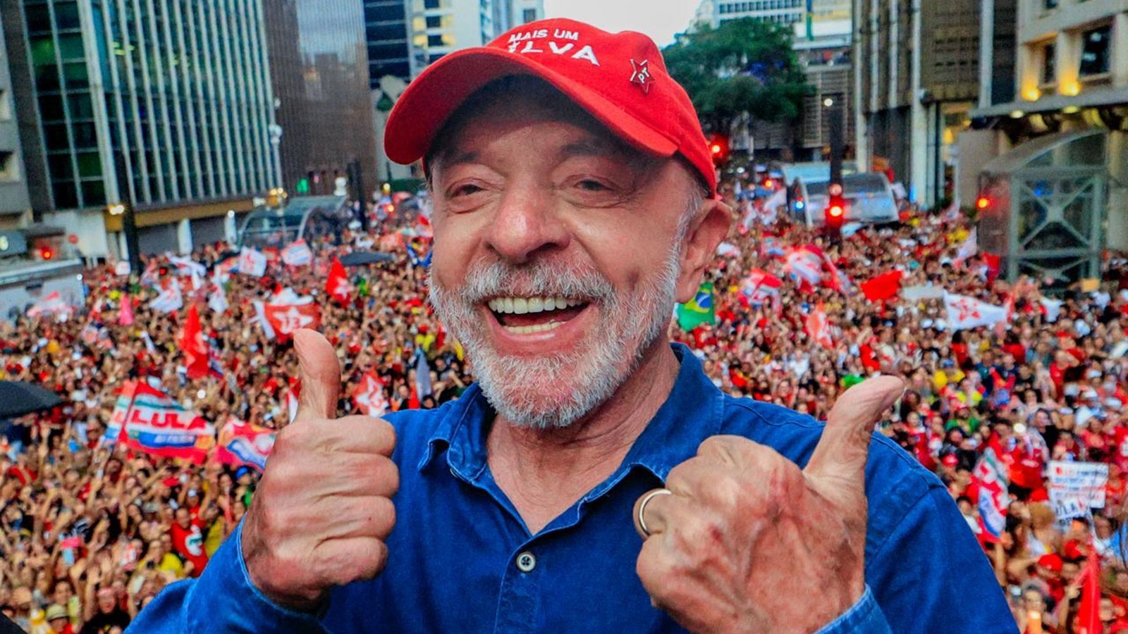 Lula sorrindo, dois polegares para cima, boné vermelho, pin PT, multidão ao fundo