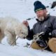 Homem barbudo com gorro Carhartt e luvas Hestra acaricia cachorro branco na neve