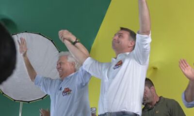 Ronaldo Caiado e Flávio Bolsonaro sorrindo, erguem os braços unidos em palco verde e amarelo