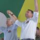 Ronaldo Caiado e Flávio Bolsonaro sorrindo, erguem os braços unidos em palco verde e amarelo