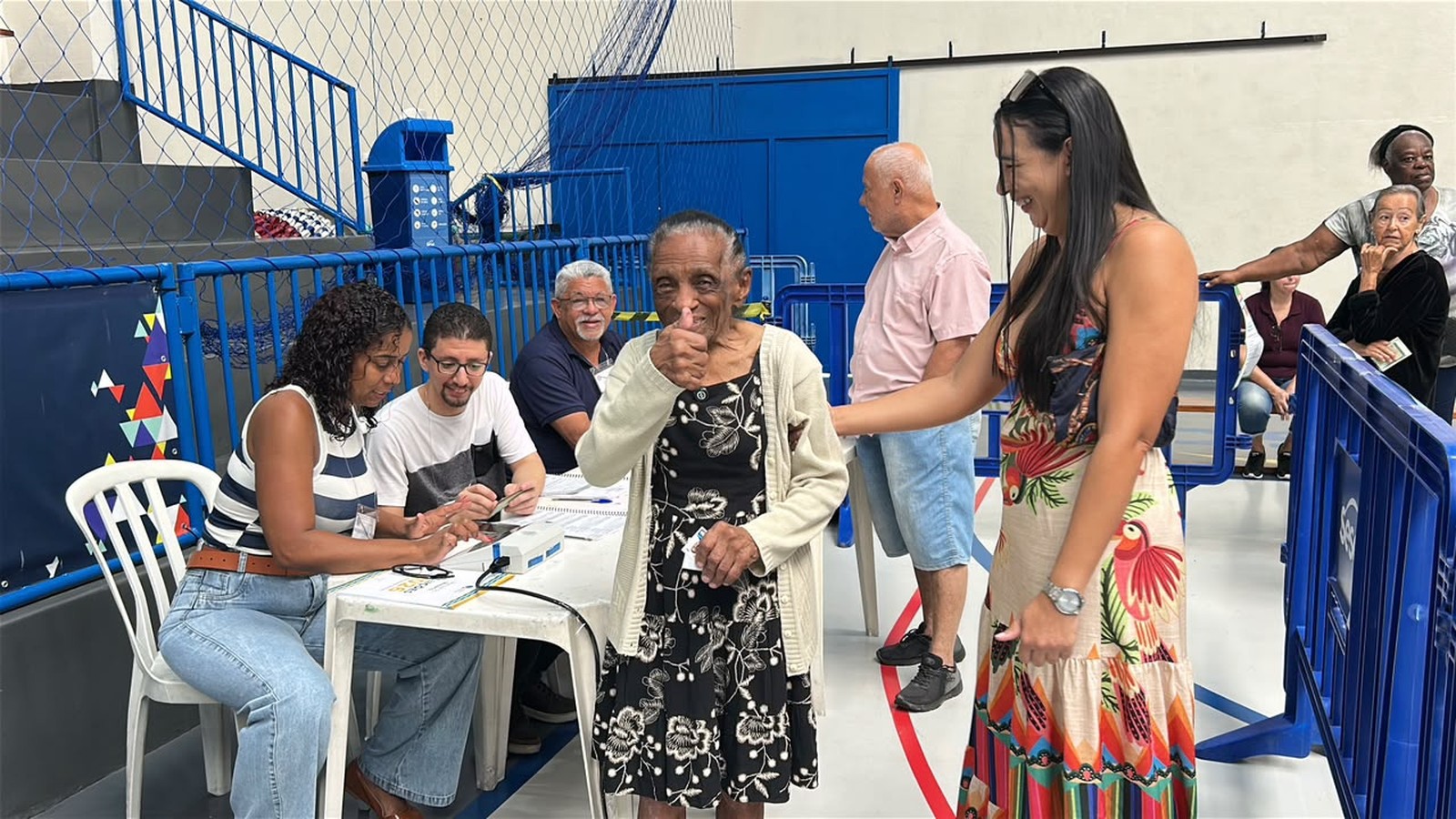 Idosa sorridente em vestido floral com polegar para cima em seção eleitoral com pessoas