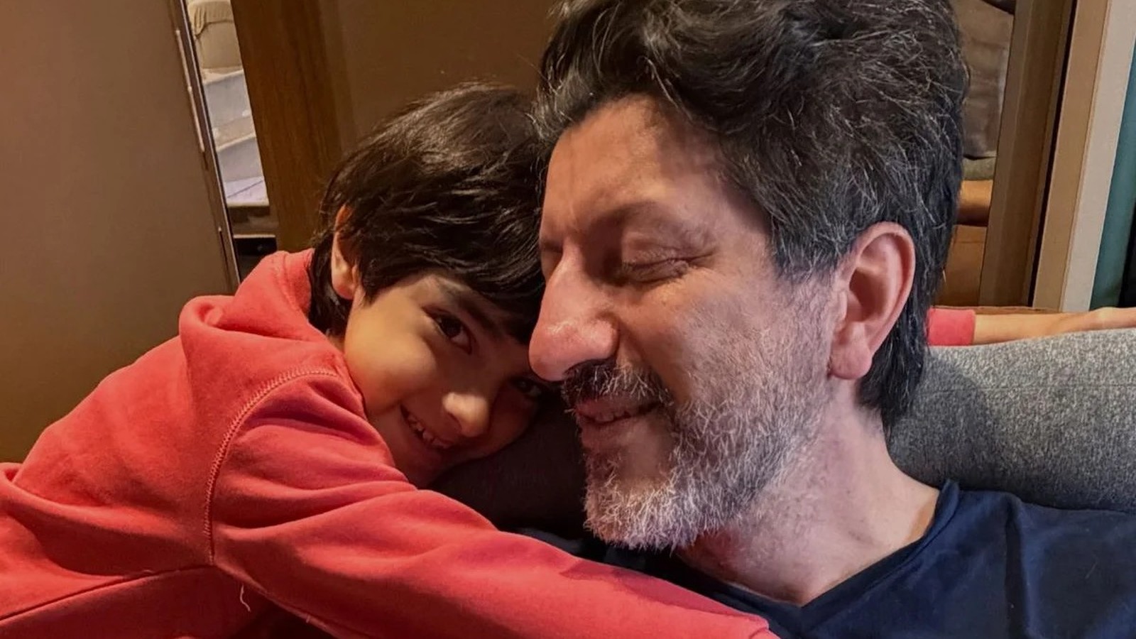 Homem de meia-idade com barba grisalha, olhos fechados, abraça menino sorridente