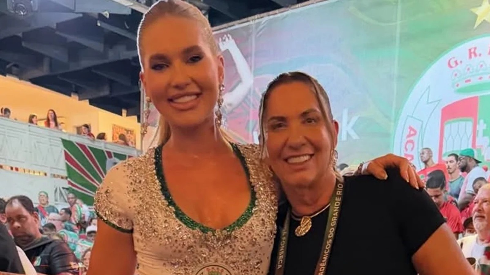 Duas mulheres sorridentes, uma loira em vestido brilhante e outra com crachá Grande Rio