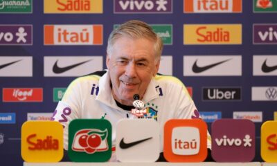 Carlo Ancelotti sorrindo em coletiva de imprensa com vários logotipos de patrocinadores