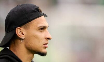 Perfil de um homem usando boné preto e brincos de cruz prateados em um ambiente desfocado