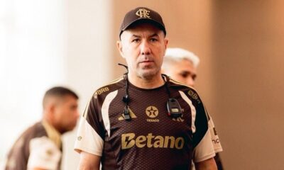 Homem com boné CRF e camisa marrom Flamengo, BRB, Texaco, Betano, fones e apito no pescoço