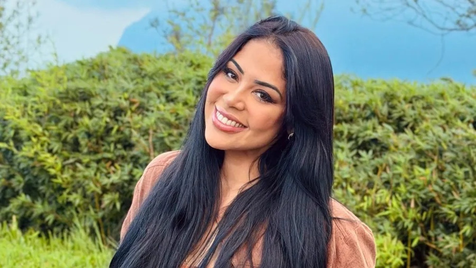 Gisele Nascimento sorri, cabelos escuros, em ambiente externo com folhagem verde