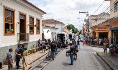 Equipe de filmagem em rua com câmera em trilhos, prédio, vans, pessoas e padaria