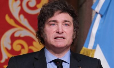 Javier Milei em terno discursa, bandeira da Argentina e parede vermelha ao fundo