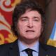 Javier Milei em terno discursa, bandeira da Argentina e parede vermelha ao fundo