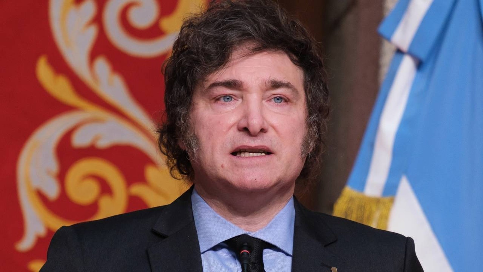 Javier Milei em terno discursa, bandeira da Argentina e parede vermelha ao fundo