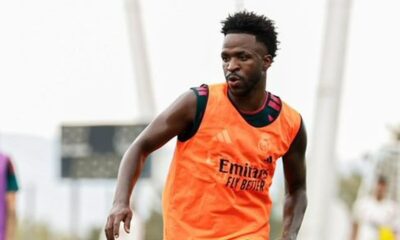Vini Jr. em campo, usando colete de treino laranja com "Emirates FLY BETTER", olhando para a direita