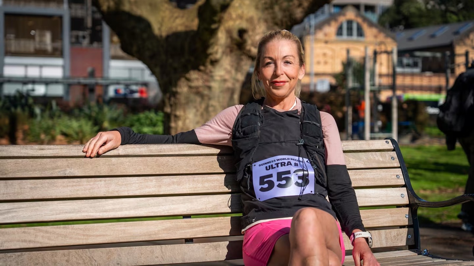Mulher sentada em banco de madeira, colete de corrida com bib "553", sorrindo