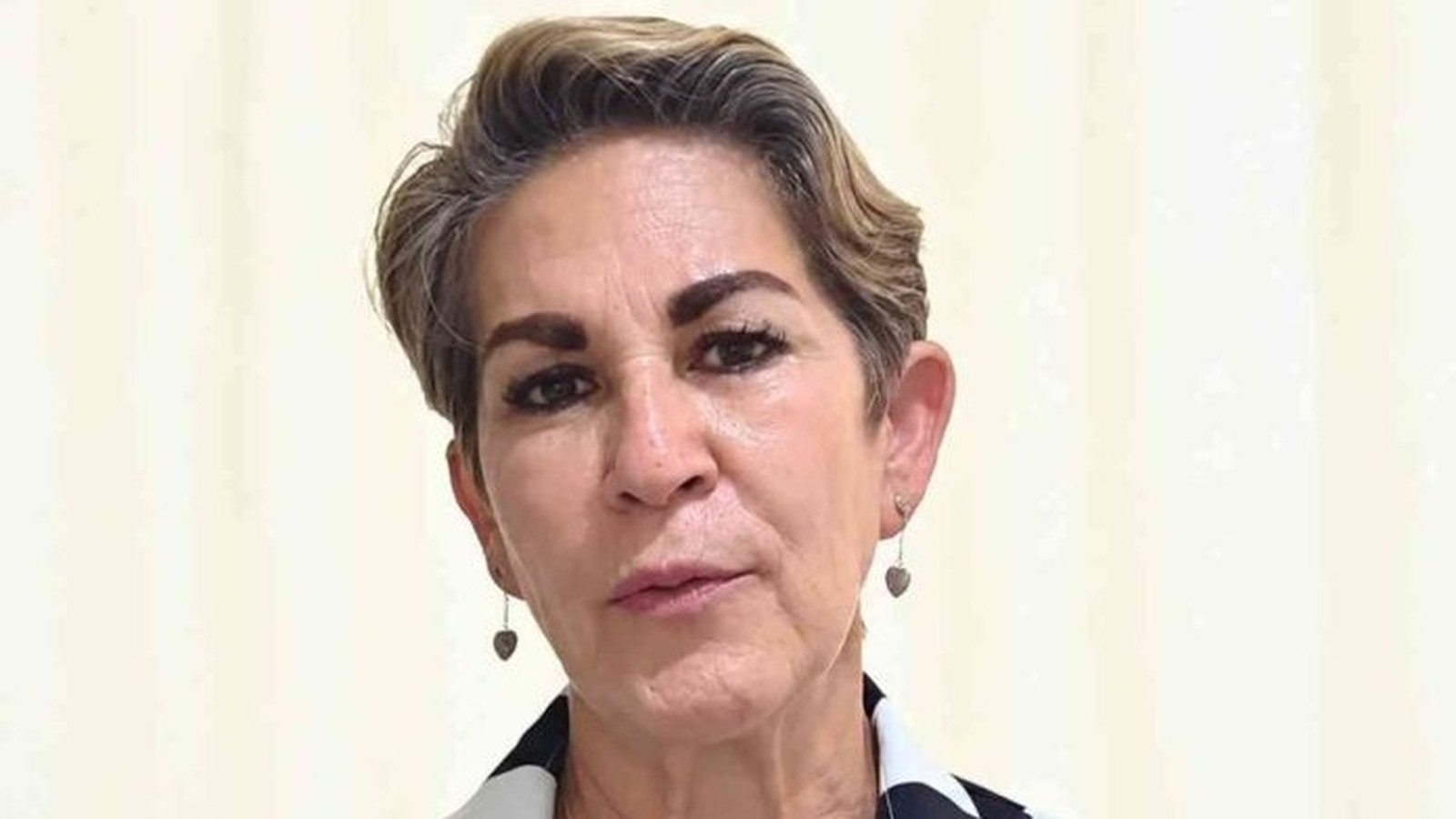 Retrato frontal de mulher madura com cabelo curto, brincos de coração e expressão neutra