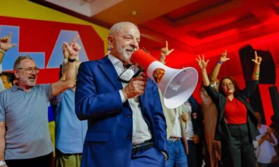 Lula sorrindo, de terno azul, segura megafone em evento de campanha com apoiadores.
