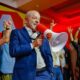 Lula sorrindo, de terno azul, segura megafone em evento de campanha com apoiadores.