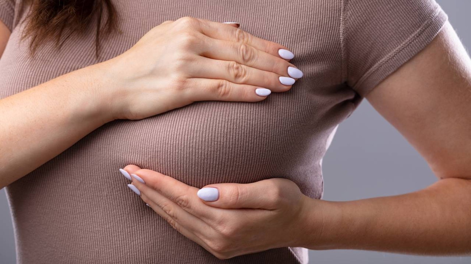 Mãos femininas com unhas claras sobre os seios, vestindo blusa marrom canelada