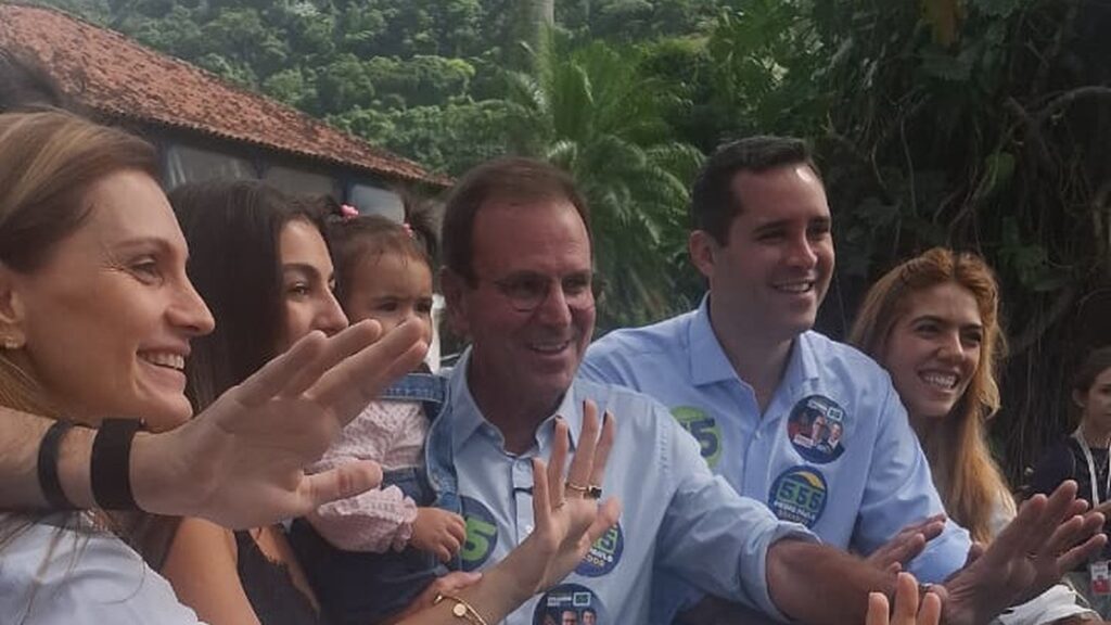 Eduardo Paes e família sorrindo, acenando, com broches de campanha '5' e '555'