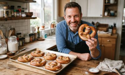 Homem sorridente na cozinha segura pretzel assado, com assadeira cheia de pretzels na bancada
