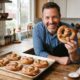 Homem sorridente na cozinha segura pretzel assado, com assadeira cheia de pretzels na bancada