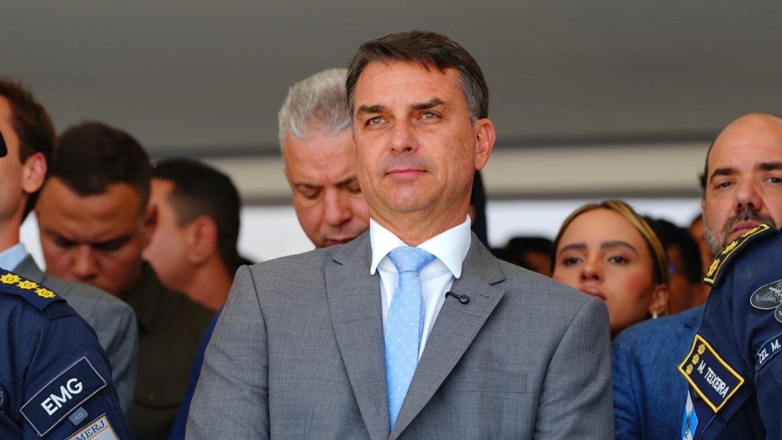 Flávio Bolsonaro de terno e gravata azul claro, cercado por outras pessoas em evento