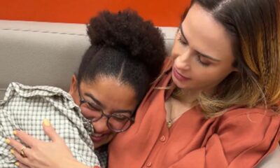 Duas mulheres se abraçam em um sofá. Uma de óculos e camisa xadrez sorri.