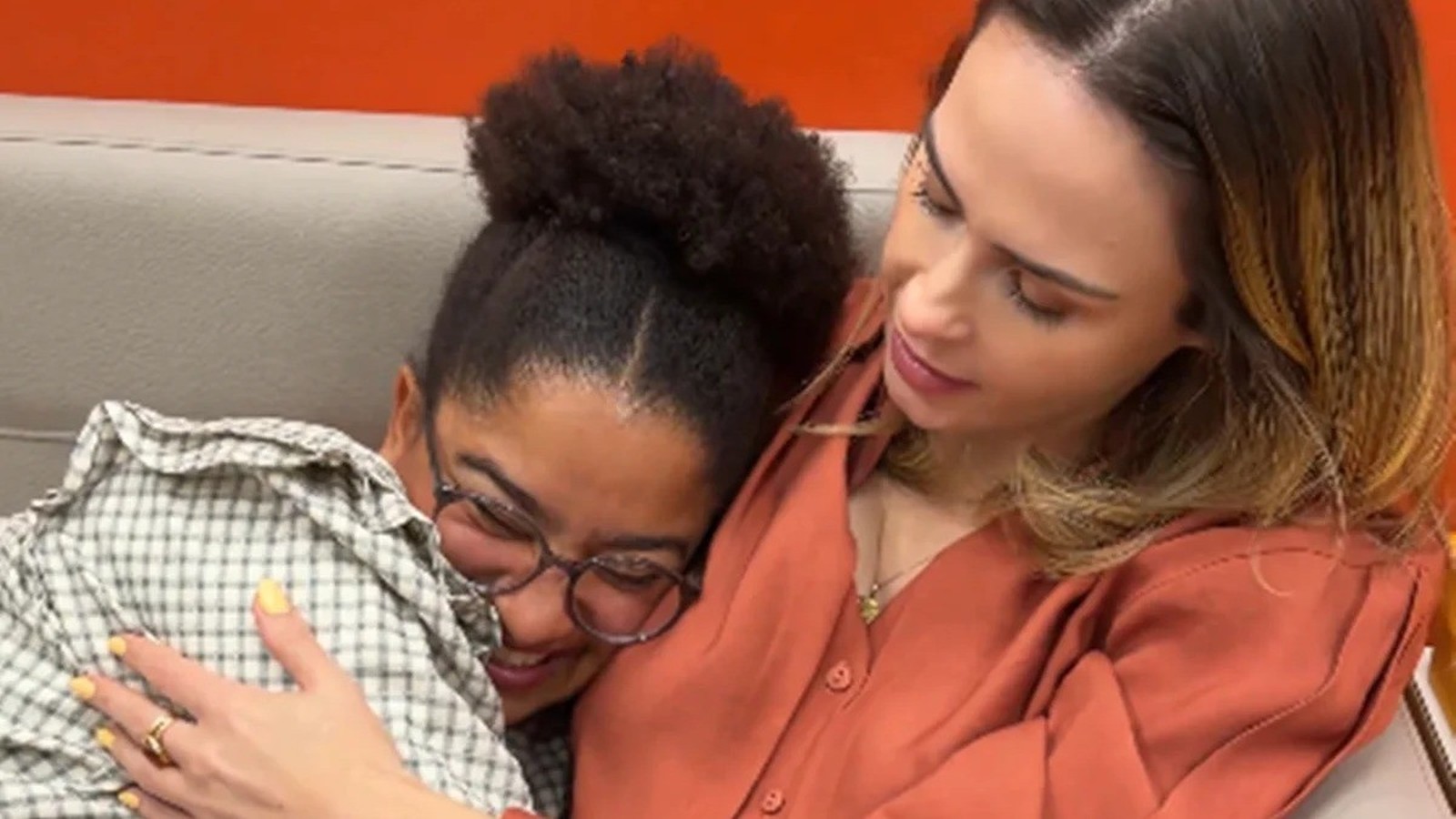 Duas mulheres se abraçam em um sofá. Uma de óculos e camisa xadrez sorri.
