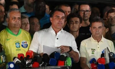 Flávio Bolsonaro, camisa branca, ladeado por dois homens em entrevista coletiva com microfones.