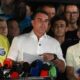 Flávio Bolsonaro, camisa branca, ladeado por dois homens em entrevista coletiva com microfones.