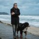 Homem de casaco, cão, caminha em passarela de madeira na praia com mar agitado