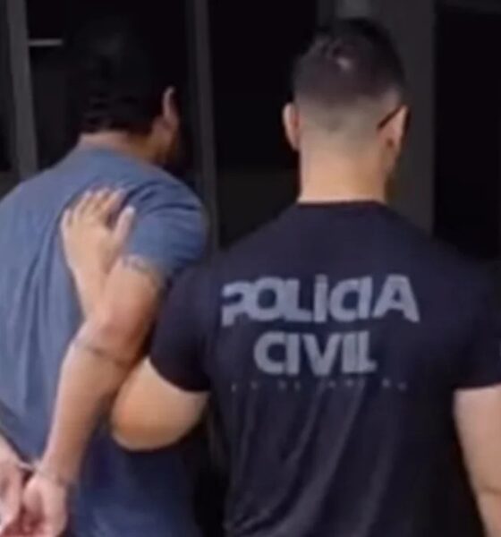 Dois homens de costas, um algemado sendo escoltado por policial de camiseta com os dizeres 'POLÍCIA CIVIL'.
