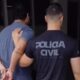 Dois homens de costas, um algemado sendo escoltado por policial de camiseta com os dizeres 'POLÍCIA CIVIL'.