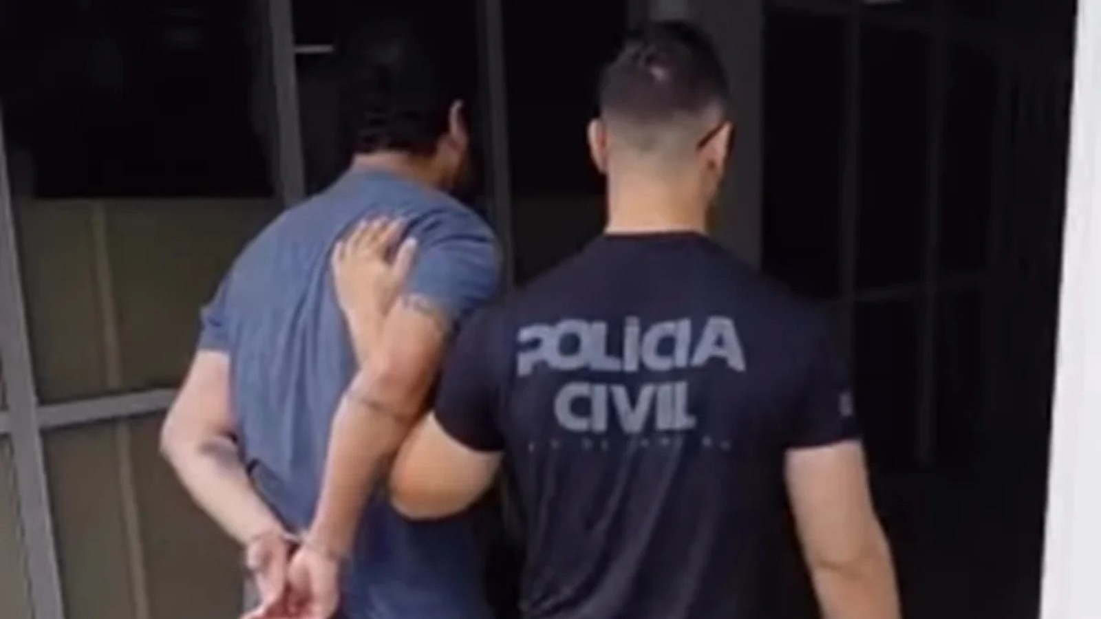 Dois homens de costas, um algemado sendo escoltado por policial de camiseta com os dizeres 'POLÍCIA CIVIL'.