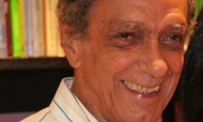 Close-up de homem idoso sorrindo, camisa listrada, cabelo grisalho e fundo borrado de livros