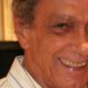 Close-up de homem idoso sorrindo, camisa listrada, cabelo grisalho e fundo borrado de livros