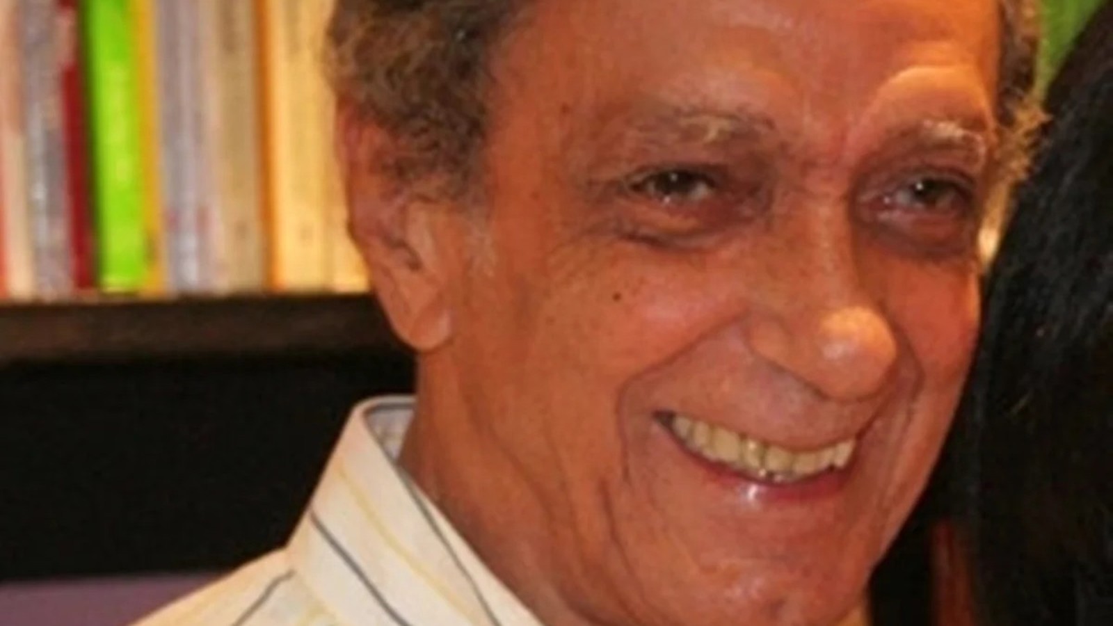 Close-up de homem idoso sorrindo, camisa listrada, cabelo grisalho e fundo borrado de livros
