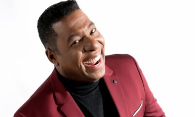 Netinho de Paula sorrindo, usando jaqueta vinho e gola alta preta, em fundo branco