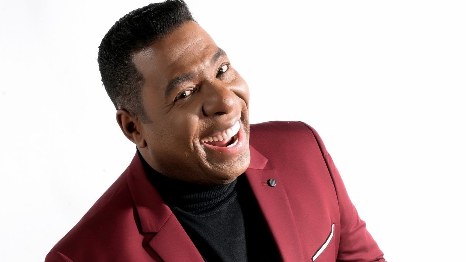 Netinho de Paula sorrindo, usando jaqueta vinho e gola alta preta, em fundo branco